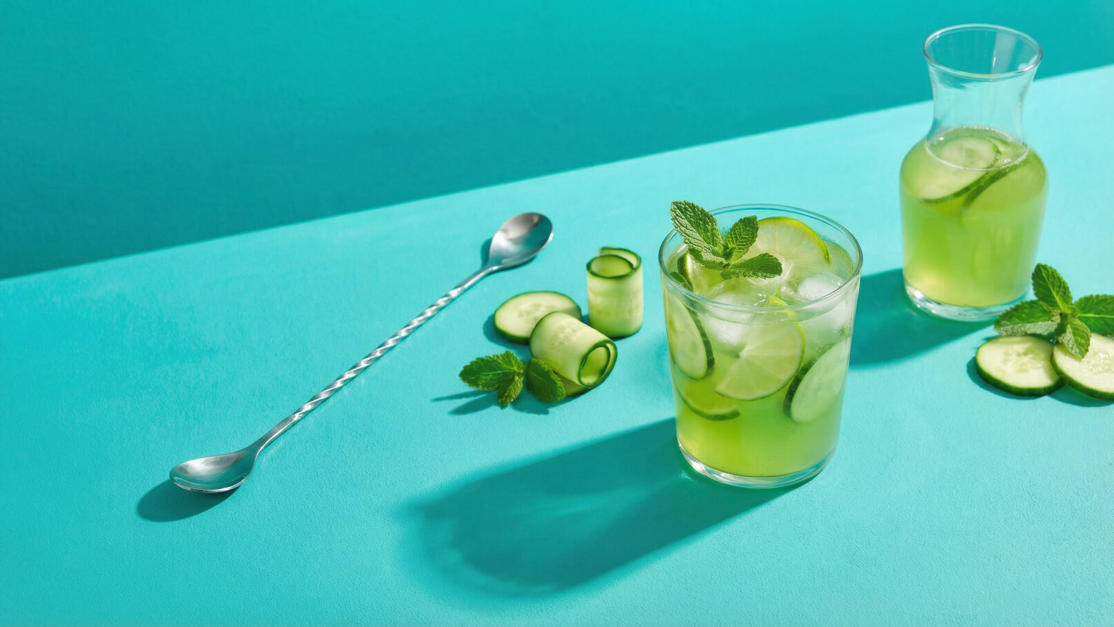 Ein Glas mit einem erfrischenden grünen Getränk, garniert mit Minze und Gurkenscheiben, steht auf einem hellblauen Tisch. Daneben ist eine Karaffe mit demselben Getränk und einige Gurkenraspel zu sehen. Eine Löffel aus Metall liegt ebenfalls auf dem Tisch.