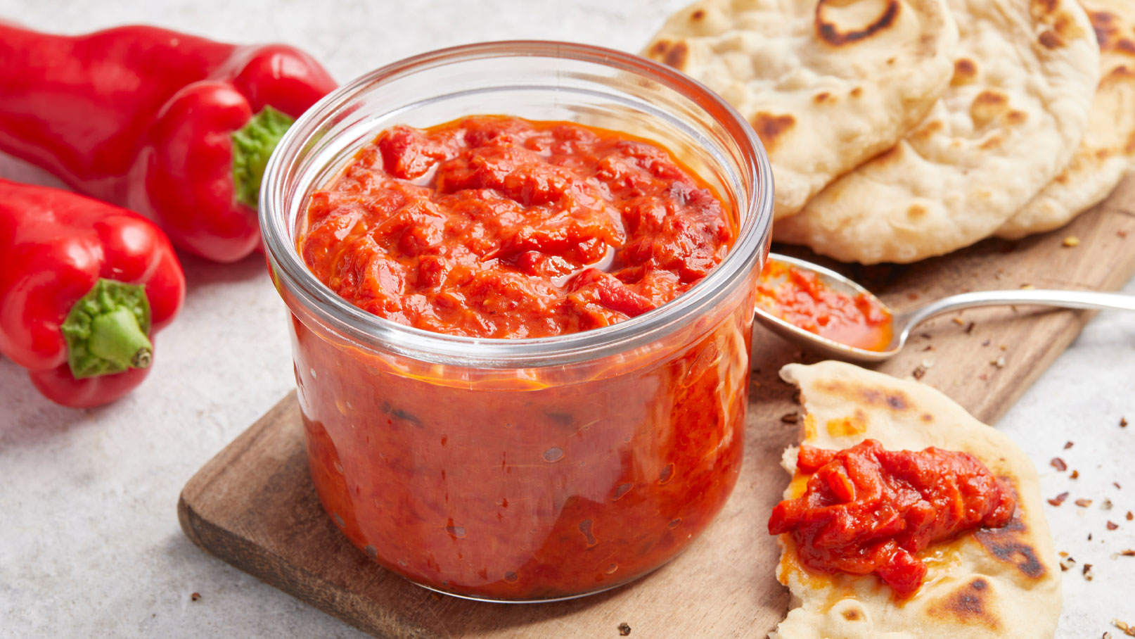 Ein Glas mit roter Sauce steht auf einem Holzbrett. Neben dem Glas liegen einige rote Paprika und ein Stück Fladenbrot, das mit der Sauce bestrichen ist. Die Sauce hat eine dicke, appetitliche Konsistenz.