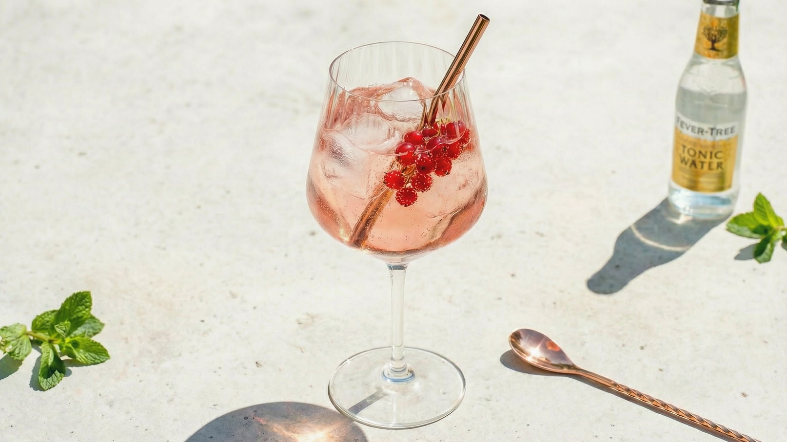 Ein elegantes Glas gefüllt mit einem rosa Getränk, garniert mit frischen roten Beeren und einem Strohhalm. Daneben steht eine Flasche Tonic Water, und im Vordergrund sind einige Minzblätter zu sehen. Der Hintergrund ist hell und neutral.