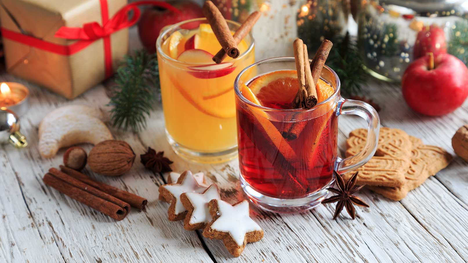 Zwei Tassen Glühwein mit Zimtstangen auf einem Holztisch, umgeben von sternförmigen Keksen, Zimtstangen, Nüssen und einer Geschenkbox.