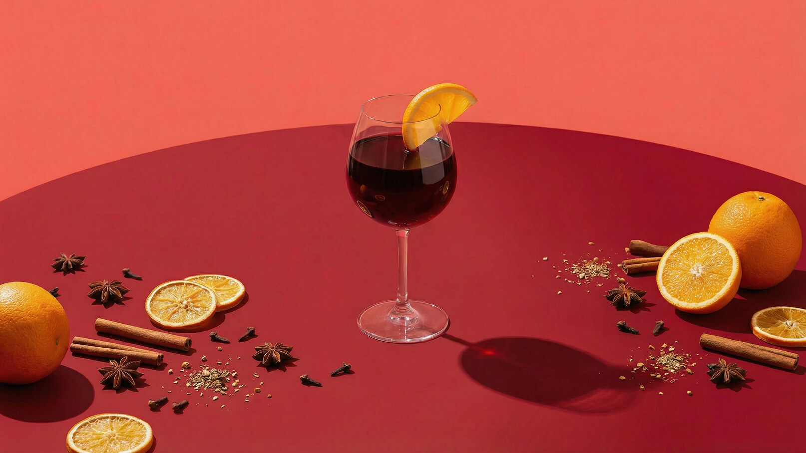 Ein Glas mit dunklem Getränk steht auf einem roten Tisch. Am Glas ist eine Orangenscheibe dekoriert. Um das Glas herum liegen ganze und geschnittene Orangen, Zimtstangen, Sternanis und Nelken, die eine warme, festliche Stimmung vermitteln.
