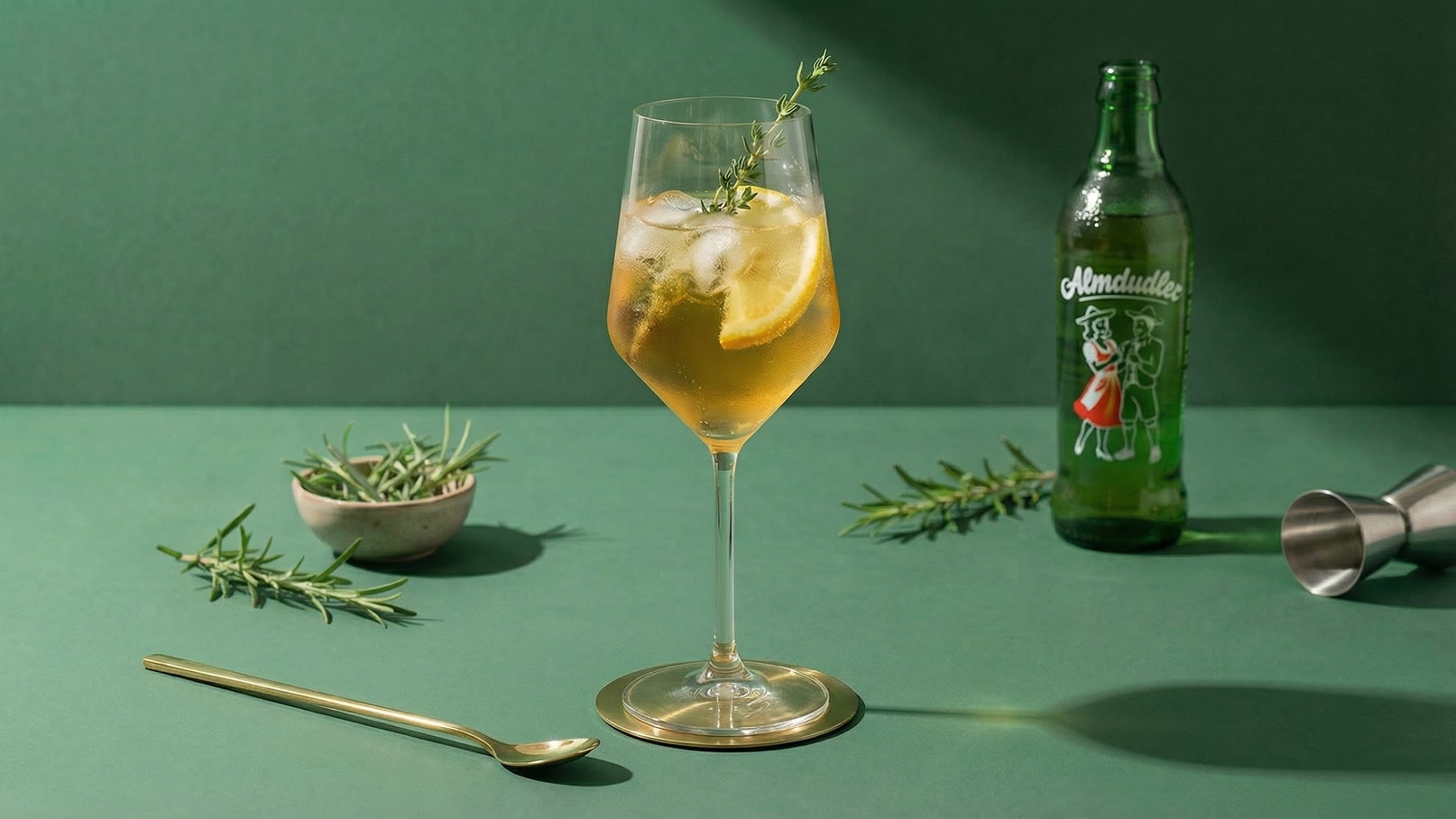 Ein elegantes Glas mit einem gelblich-braunen Getränk, Eiswürfeln und einer Zitronenscheibe. Darüber ist ein Zweig Thymian dekoriert. Neben dem Glas steht eine grüne Flasche mit dem Aufdruck "Almdudler" und ein kleiner Behälter mit frischen Kräutern. Der Hintergrund ist grün.