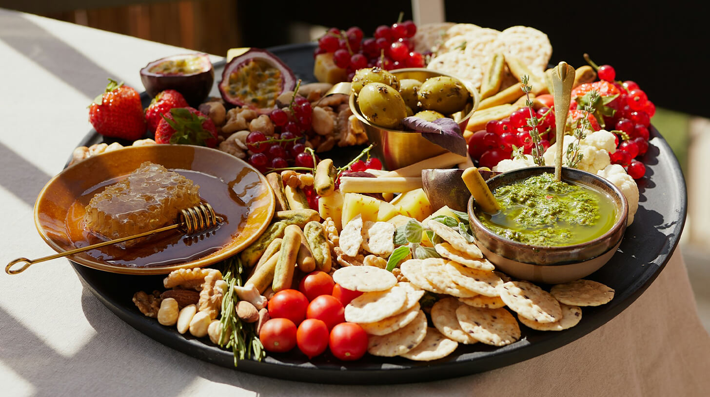 Eine Platte mit Crackern, grünem Dip, Honigwabe, Kirschtomaten, Weintrauben, Passionsfrüchten, Erdbeeren, Nüssen und gefüllten Oliven.