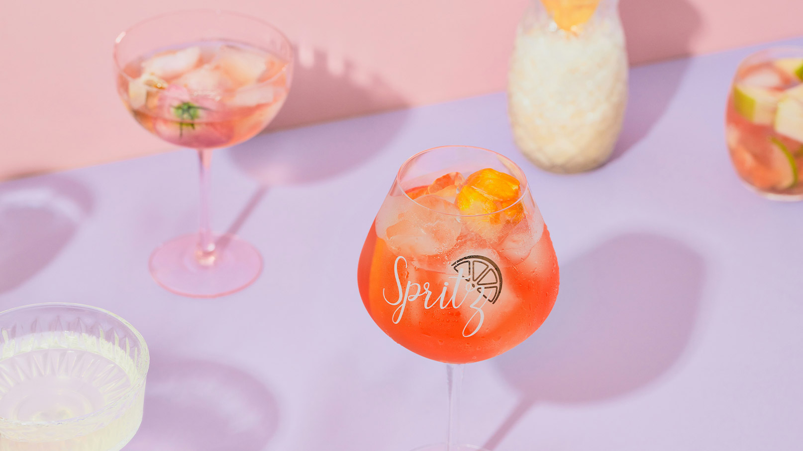 Ein großes Glas mit rotem Cocktail und Eis, beschriftet mit „Spritz“. Im Hintergrund sind weitere stilvolle Cocktails zu sehen, die auf einem lilafarbenen Tisch stehen. Die Atmosphäre ist fröhlich und einladend.