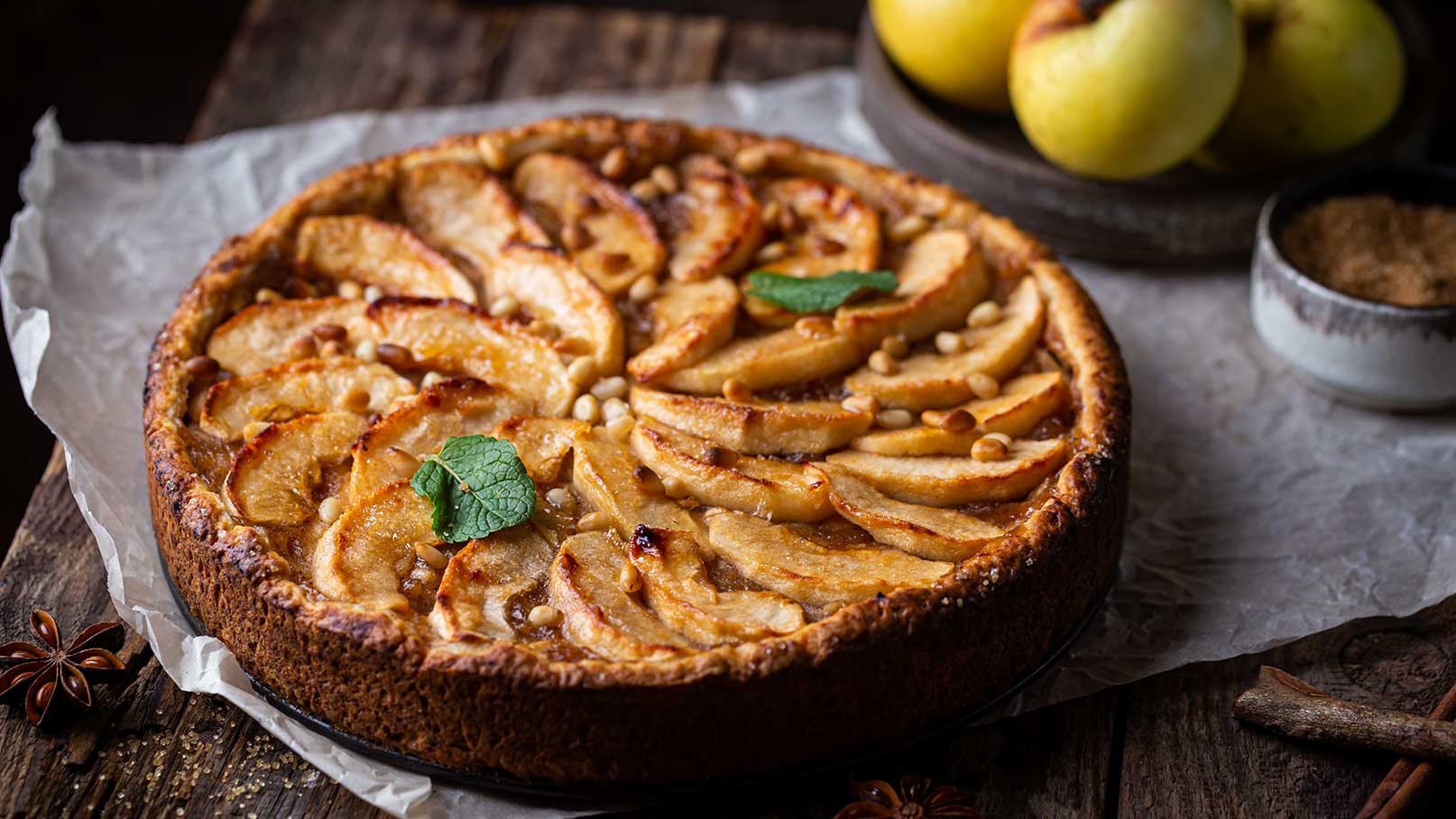 Apfelkuchen mit Apfelscheiben und Minzblättern auf einer Holzoberfläche. Im Hintergrund stehen eine Schale mit frischen Äpfeln.