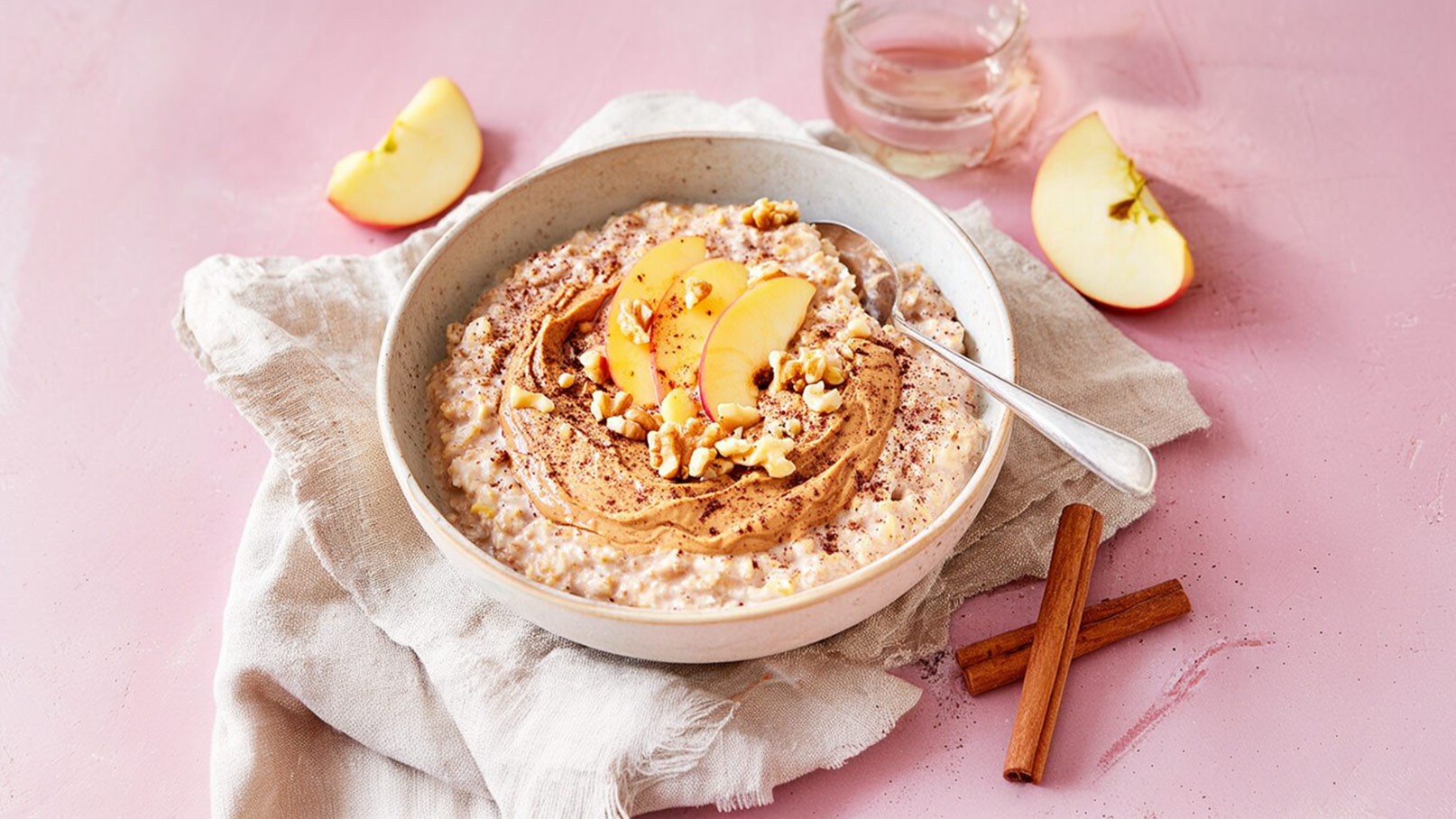 Eine Schale Haferbrei mit
Apfelscheiben, Nüssen und einer cremigen Erdnussbutter-Schicht. Der Brei wird mit Zimt bestreut. Neben der Schale liegen einige Apfelschnitze und Zimtstangen auf einem hellen Tuch. Der Hintergrund ist rosa.