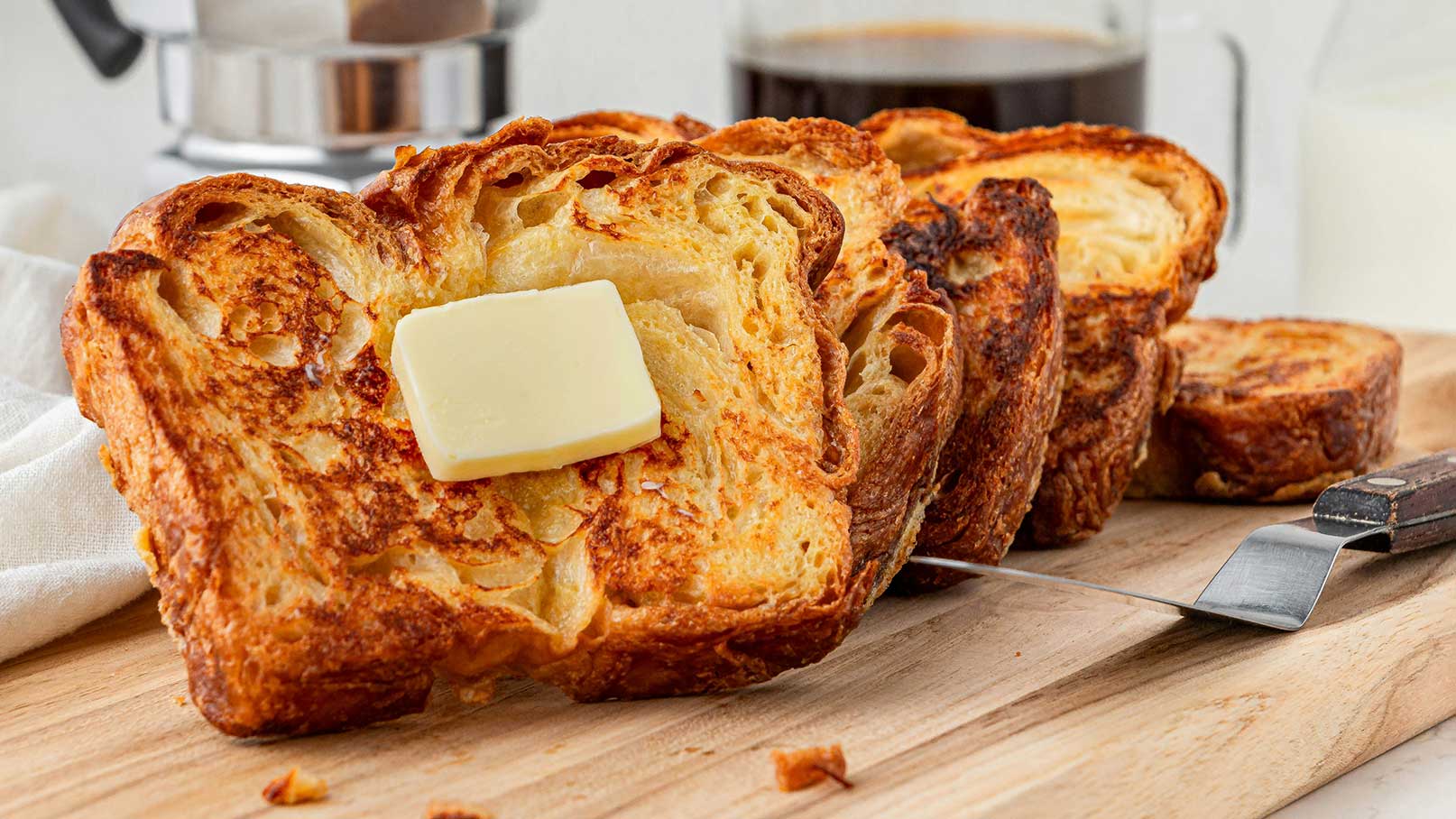 Drei goldbraun gebratene, geschnittene Brotscheiben liegen auf einem Holzbrett. Auf einer Scheibe befindet sich ein Stück Butter. Im Hintergrund sind eine Kaffeemaschine und ein Glas Milch zu sehen.