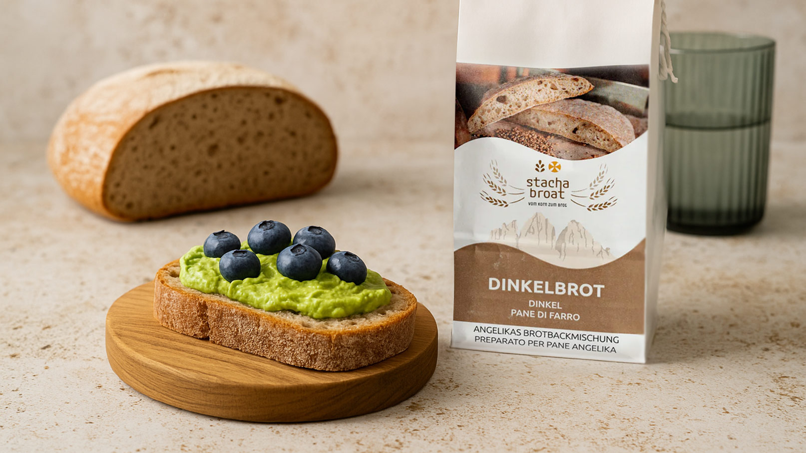 Ein Stück Dinkelbrot liegt auf einem Holzbrett. Darauf befindet sich eine Schicht grüner Avocadocreme, die mit drei Heidelbeeren dekoriert ist. Im Hintergrund steht eine Verpackung für Dinkelbrot und ein Glas.