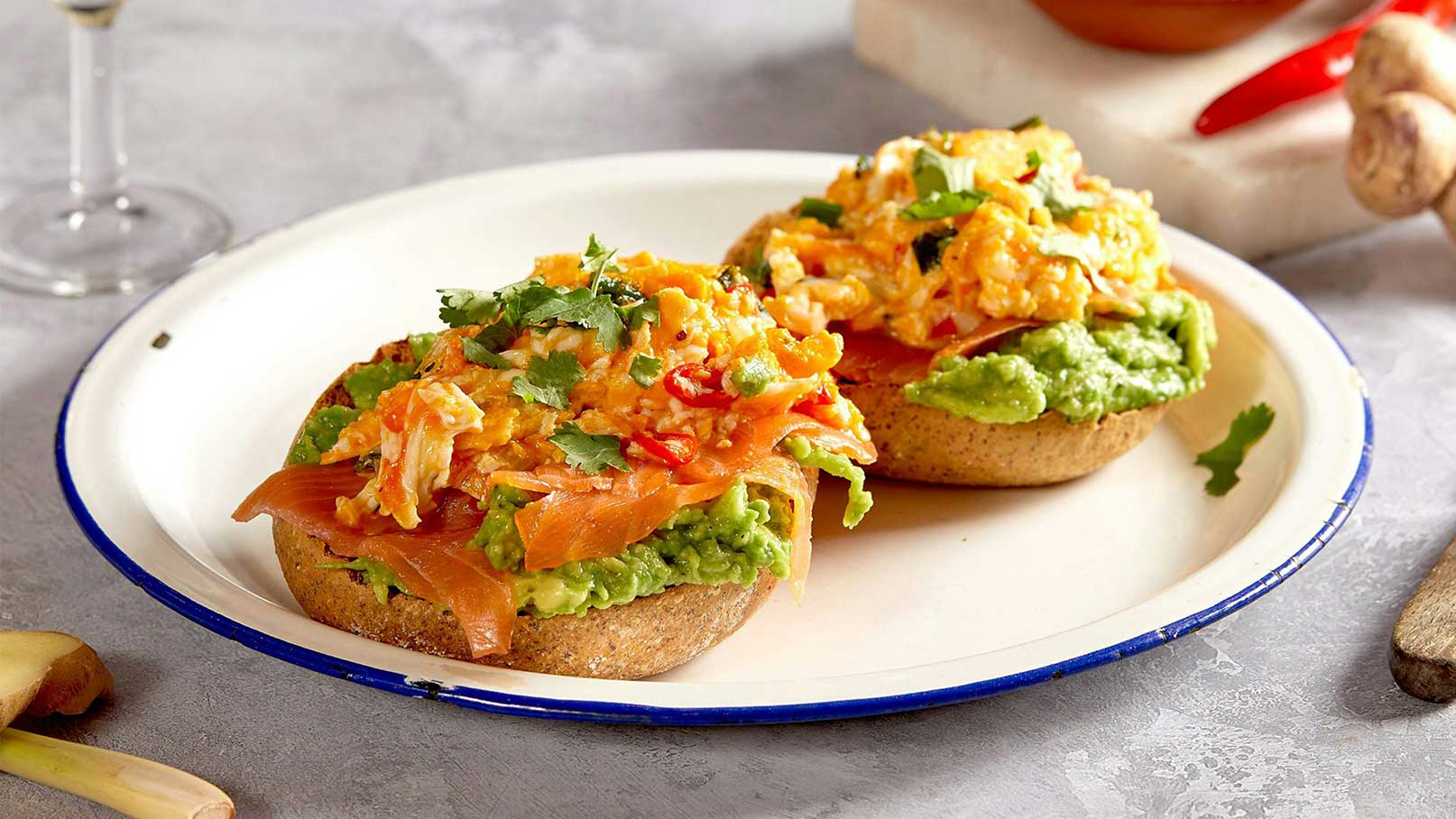 Zwei belegte Brötchen auf einem weißen Teller. Jedes Brötchen ist mit einer Schicht Guacamole, geräuchertem Lachs und einem bunten Rührei, das mit frischen Kräutern und Chili garniert ist, belegt. Der Teller liegt auf einer grauen Oberfläche.