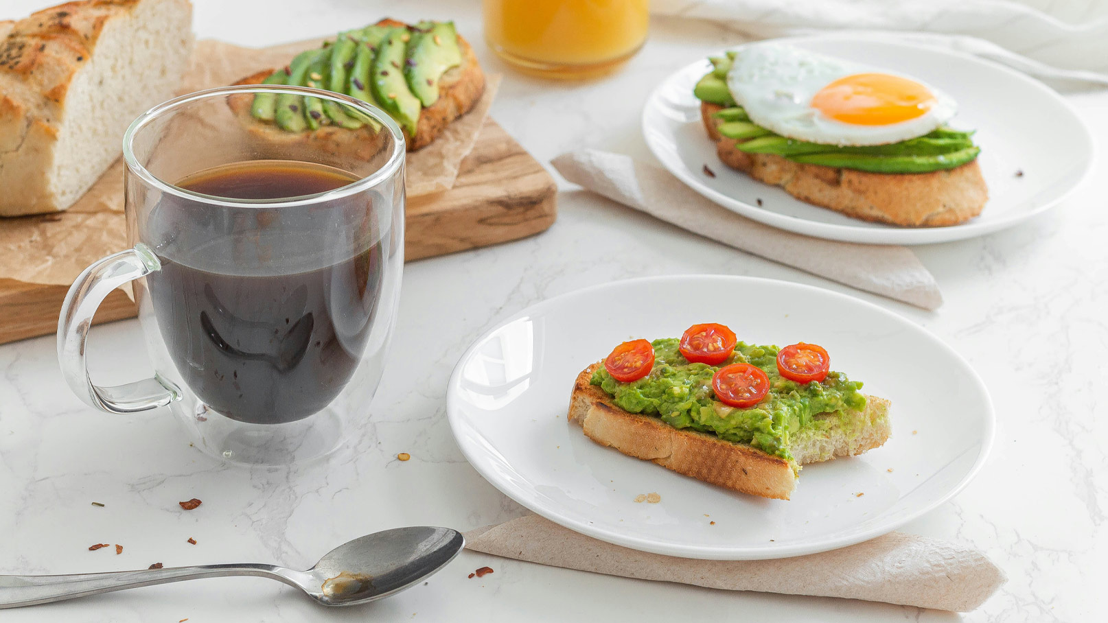 Auf dem Tisch steht eine dampfende Tasse schwarzer Kaffee. Nebenbei liegt eine Scheibe Toast mit Guacamole und Cherrytomaten. Im Hintergrund sind weitere Brotscheiben mit Avocado und ein Frühstücksei zu sehen. Die Atmosphäre wirkt einladend und gemütlich.