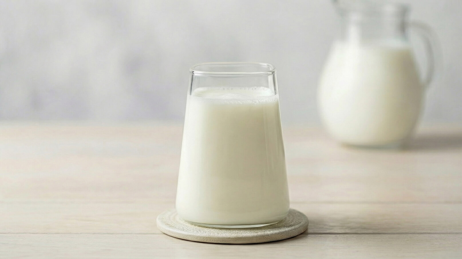 Ein Glas Milch steht auf einem Untersetzer. Im Hintergrund ist eine Kanne mit Milch zu sehen. Die Umgebung wirkt neutral und hell, was die Frische des Milchprodukts unterstreicht.