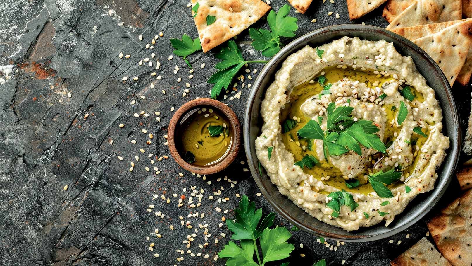 Eine Schale mit Hummus ist mit Olivenöl und frischem Grün garniert, umgeben von Sesam und frischem Brot auf einer dunklen Oberfläche. Eine kleine Schale mit Dip steht neben der Hauptschale.