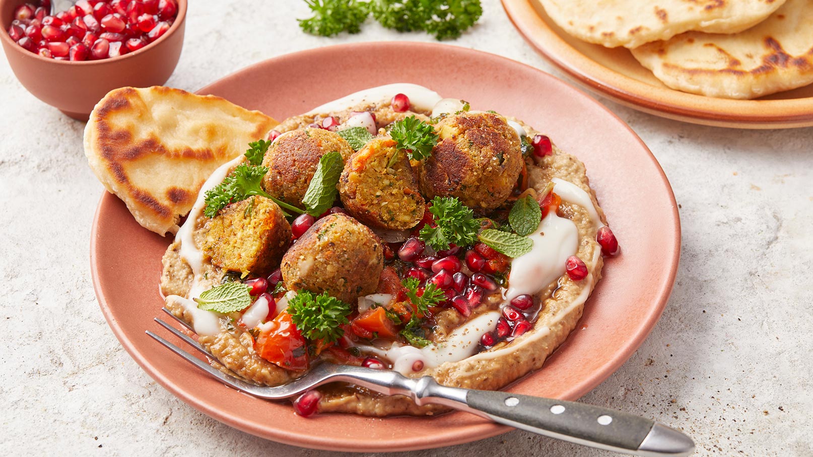 Ein Teller mit Falafel, garniert mit frischem Gemüse, Joghurt und Granatapfelkernen, auf einer Basis aus pürierten Hülsenfrüchten. Daneben liegt ein Stück Fladenbrot. Im Hintergrund sind weitere Fladenbrote sichtbar.
