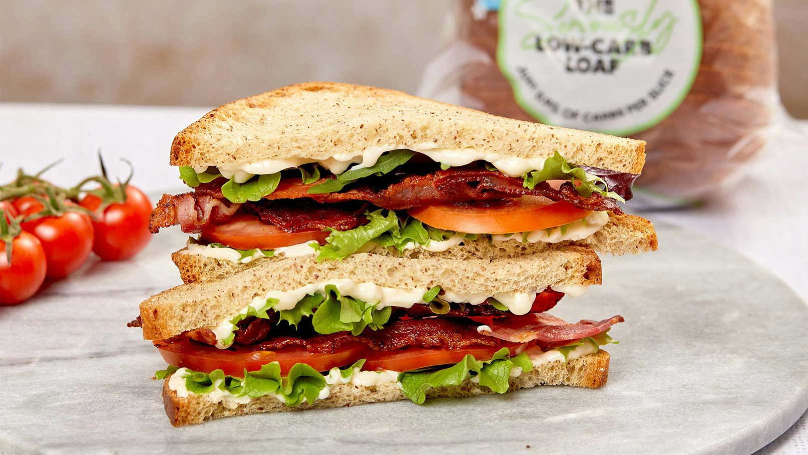 Ein hoher Sandwich-Haufen aus Vollkornbrot, gefüllt mit frischem Salat, Tomatenscheiben, knusprigem Speck und einer Cremesauce. Daneben liegen einige frische Tomaten. Das Sandwich ist auf einem Steinbrett platziert.