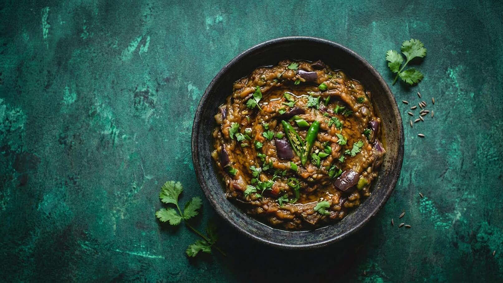 Eine dunkle Schüssel mit einer würzigen, pürierten Mischung aus Auberginen, dekoriert mit frischem Koriander und einigen grünen Chili-Streifen. Im Hintergrund sind einige Korianderblätter und Gewürze zu sehen. Der Hintergrund ist in einem tiefen Grünton gehalten.