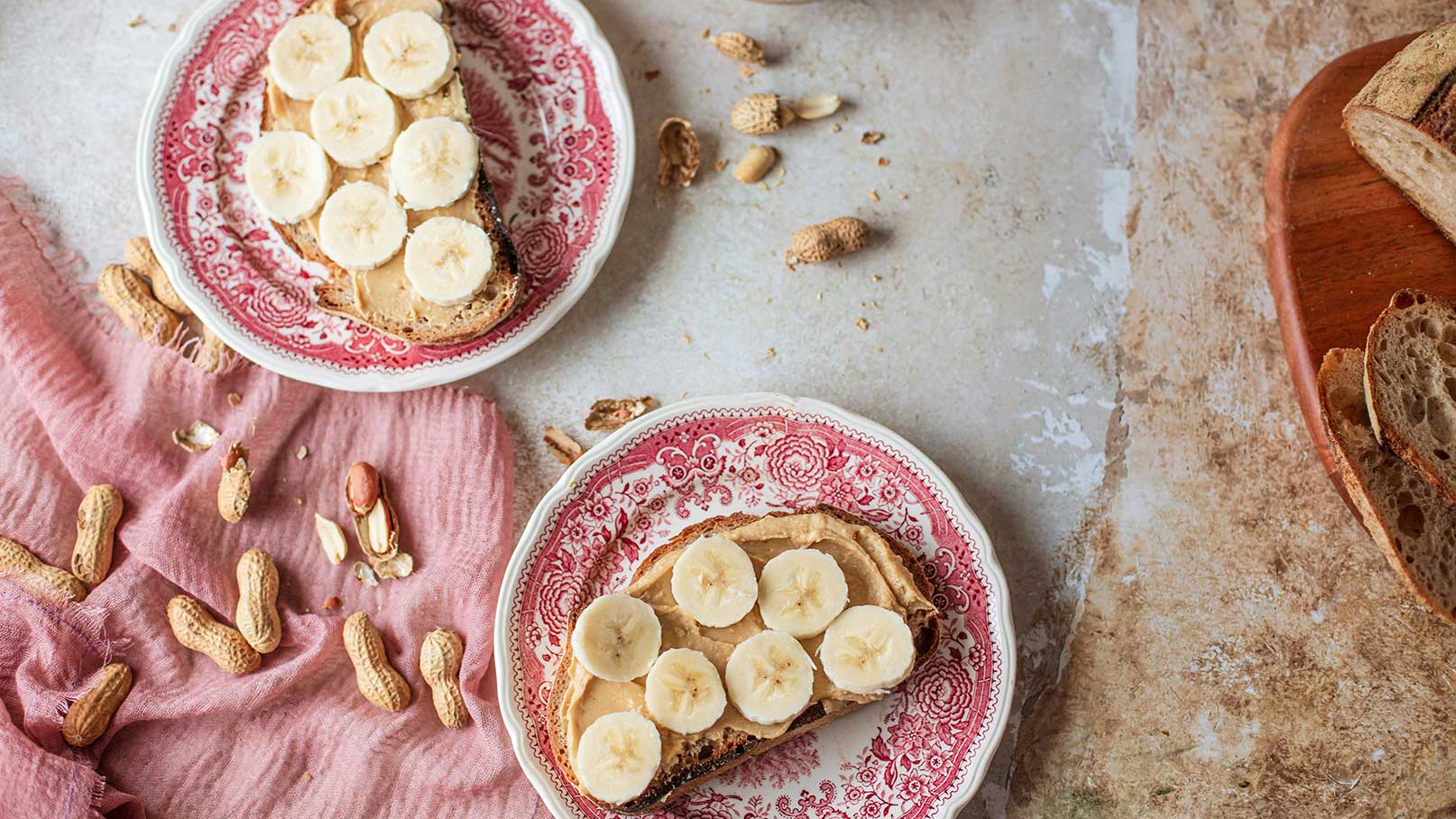 Zwei Teller mit Toast, bestrichen mit Erdnussbutter und belegt mit geschnittenen Bananen. Rund um die Teller liegen einige Erdnüsse und ein rosa Tuch. Die Teller haben einen dekorativen Rand und der Hintergrund ist in sanften, neutralen Farben gehalten.