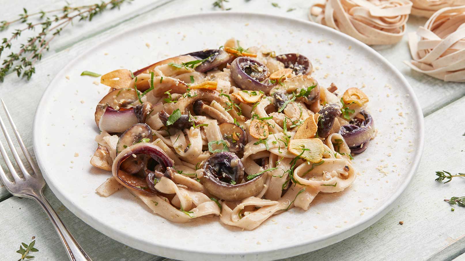 Ein Teller mit frisch zubereiteten Fettuccine, garniert mit Zwiebeln, Kräutern und einer schmackhaften Sauce. Die Pasta ist goldbraun, und einige Kräuterblätter sind darüber streut. Im Hintergrund liegen ungekochte Fettuccine.