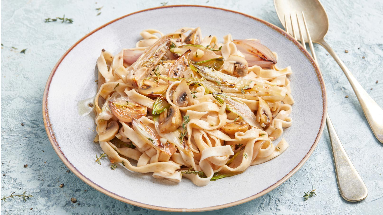 Ein Teller Pasta mit cremiger Soße, garniert mit karamellisierten Zwiebeln und Champignons. Die Nudeln sind dünn und leicht glänzend, umgeben von frischen Kräutern. Der Hintergrund ist hellblau, was den Fokus auf das Gericht lenkt.