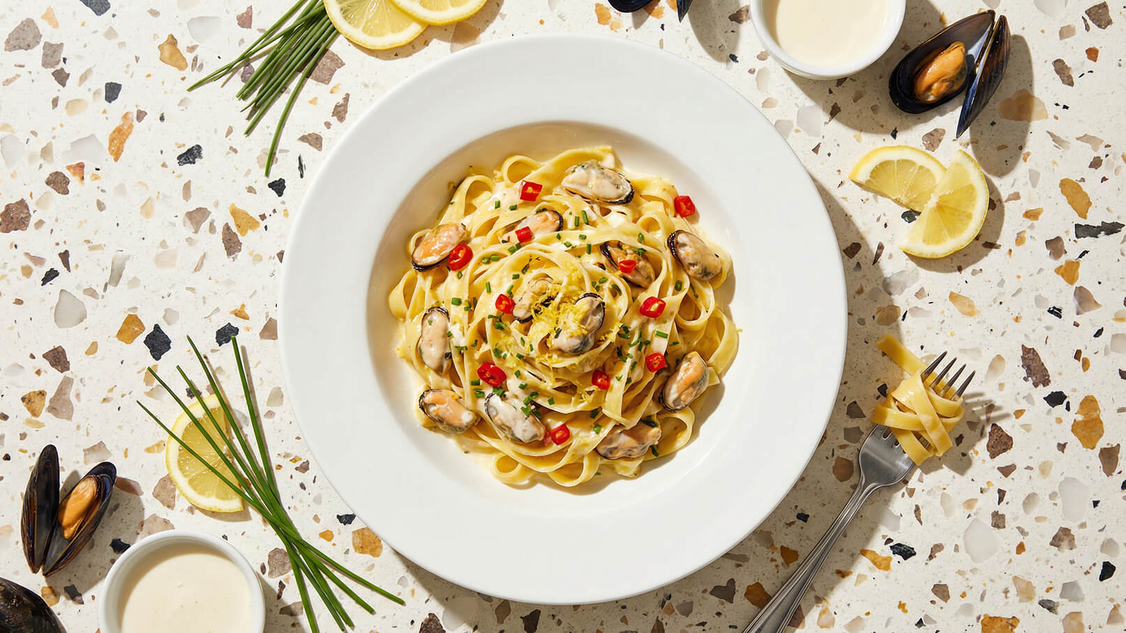 Ein Teller mit frischen Fettuccine, garniert mit Muscheln, roten Chiliflocken und frischem Schnittlauch. Neben dem Teller liegt ein kleiner Schälchen mit einer cremigen Sauce und eine halbe Zitrone. Die Bildhintergrund ist eine helle, strukturierte Oberfläche.