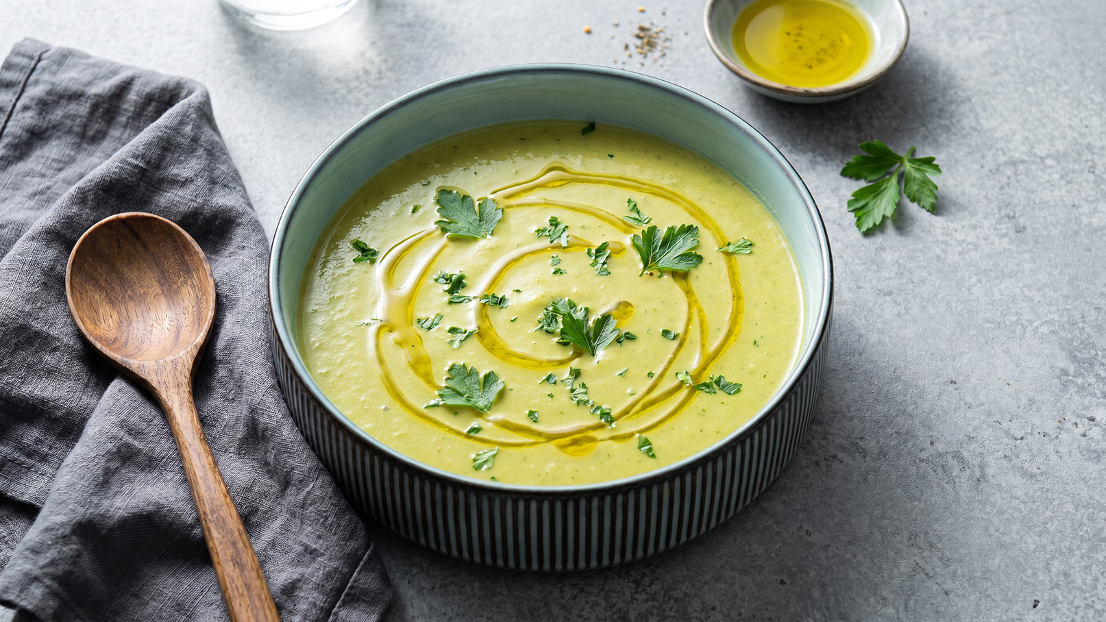 Eine Schüssel mit cremiger Suppe, garniert mit frischen Kräutern und einem Schuss Olivenöl. Neben der Schüssel liegt ein Holzlöffel und ein graues Tuch. Der Hintergrund ist schlicht und unauffällig.