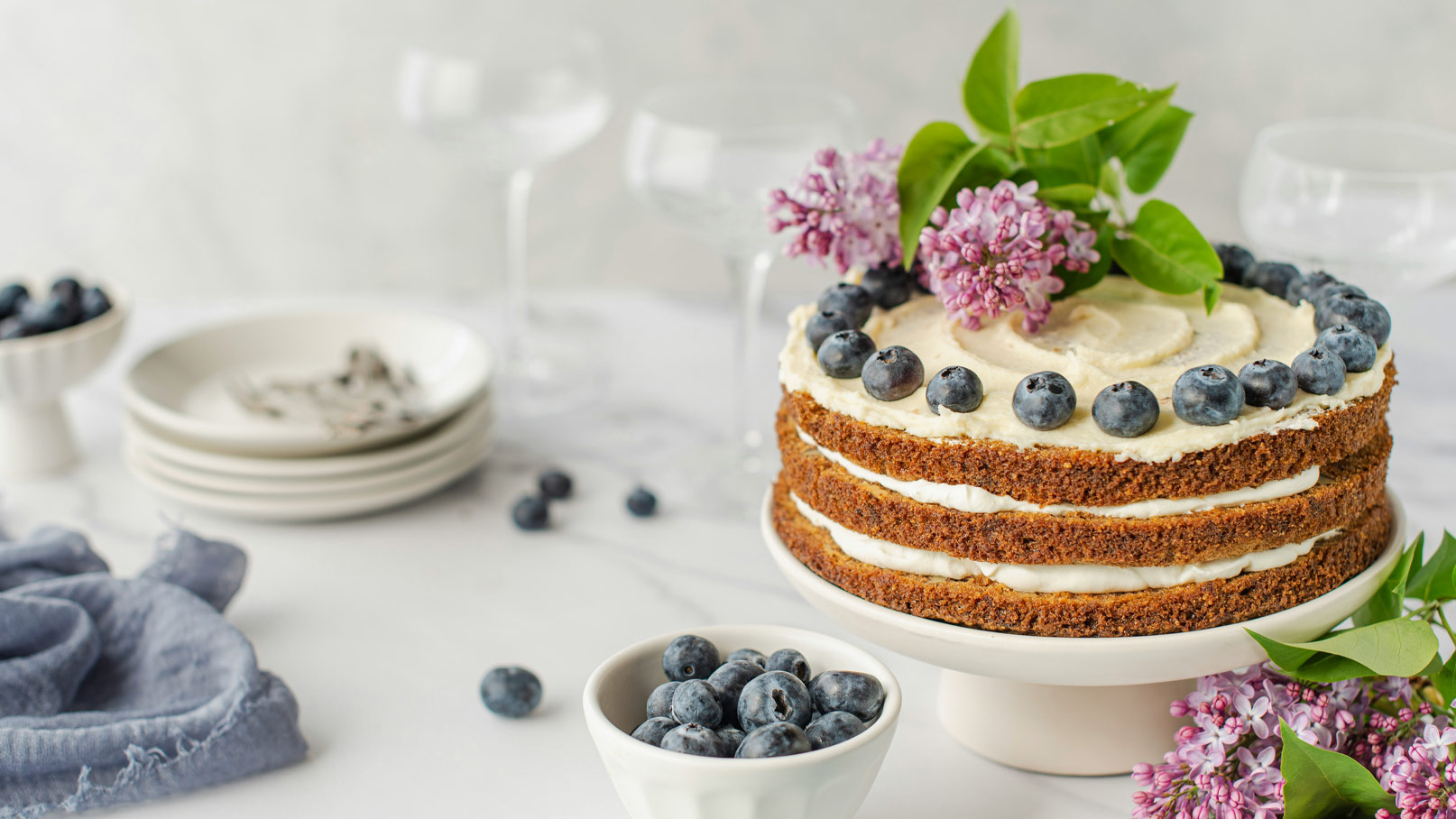 Ein dreischichtiger Kuchen auf einem weißen Teller, üppig garniert mit Blaubeeren und frischen Lilienblüten. Daneben befinden sich eine Schale mit weiteren Blaubeeren und einige Teller. Der Hintergrund ist hell und neutal gehalten.