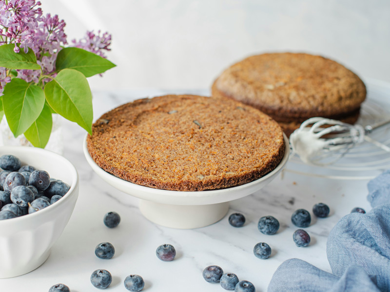 Ein Kuchen auf einer weißen Tortenplatte, umgeben von Blaubeeren auf dem Tisch. Im Hintergrund sind zwei weitere Kuchen und einige blaue Beeren verstreut. Violette Blumen und grüne Blätter ergänzen die dekorative Anordnung.