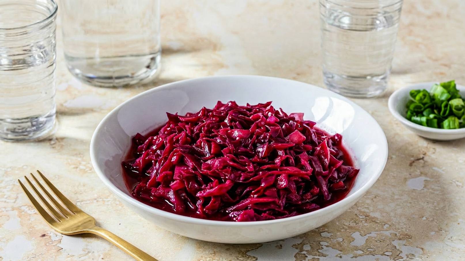 Eine weiße Schüssel mit rotviolettem, gehacktem Rotkohl steht auf einem marmorierten Tisch. Neben der Schüssel liegt eine goldene Gabel. Im Hintergrund sind Gläser mit Wasser sichtbar sowie eine kleine Schale mit gehackten Frühlingszwiebeln.