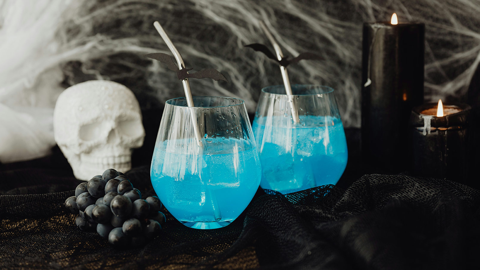 Zwei Glasbecher mit leuchtend blauem Getränk und Eiswürfeln stehen auf einem dunklen Tisch. Die Becher sind mit schwarzen Papierfledermäusen dekoriert. Daneben liegen schwarze Trauben und brennende schwarze Kerzen im Hintergrund, die eine mystische Atmosphäre erzeugen.