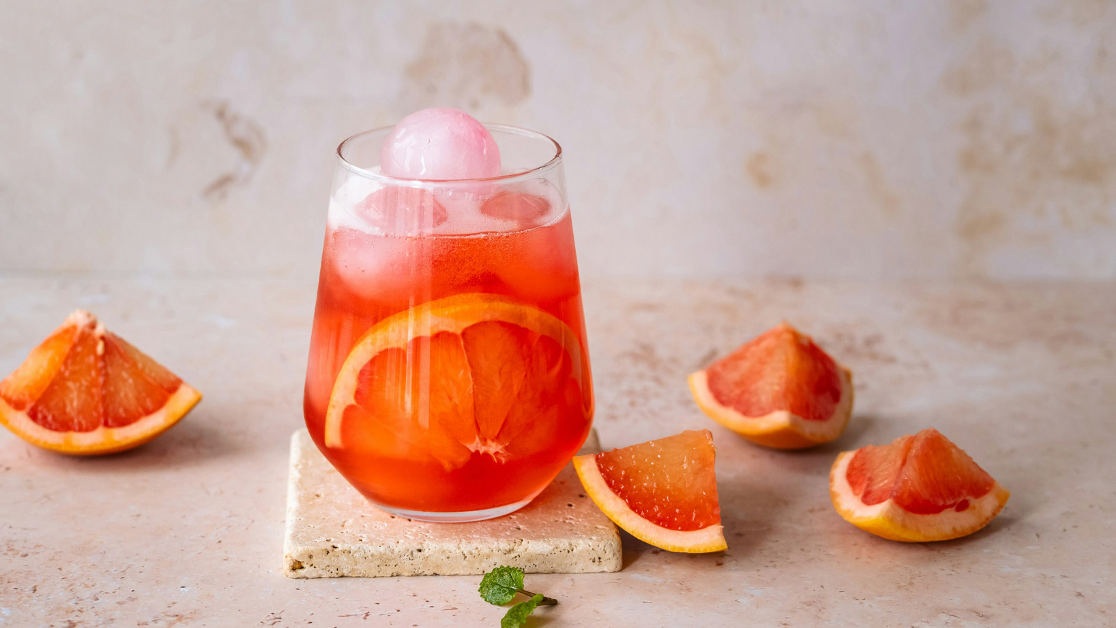 Ein Glas mit einem rosa Getränk, das Eiswürfel und eine Kugel gefrorenes Sorbet enthält. Rund um das Glas sind Fruchtscheiben von Grapefruit verteilt, die frisch und saftig wirken. Der Hintergrund ist hell und neutral.