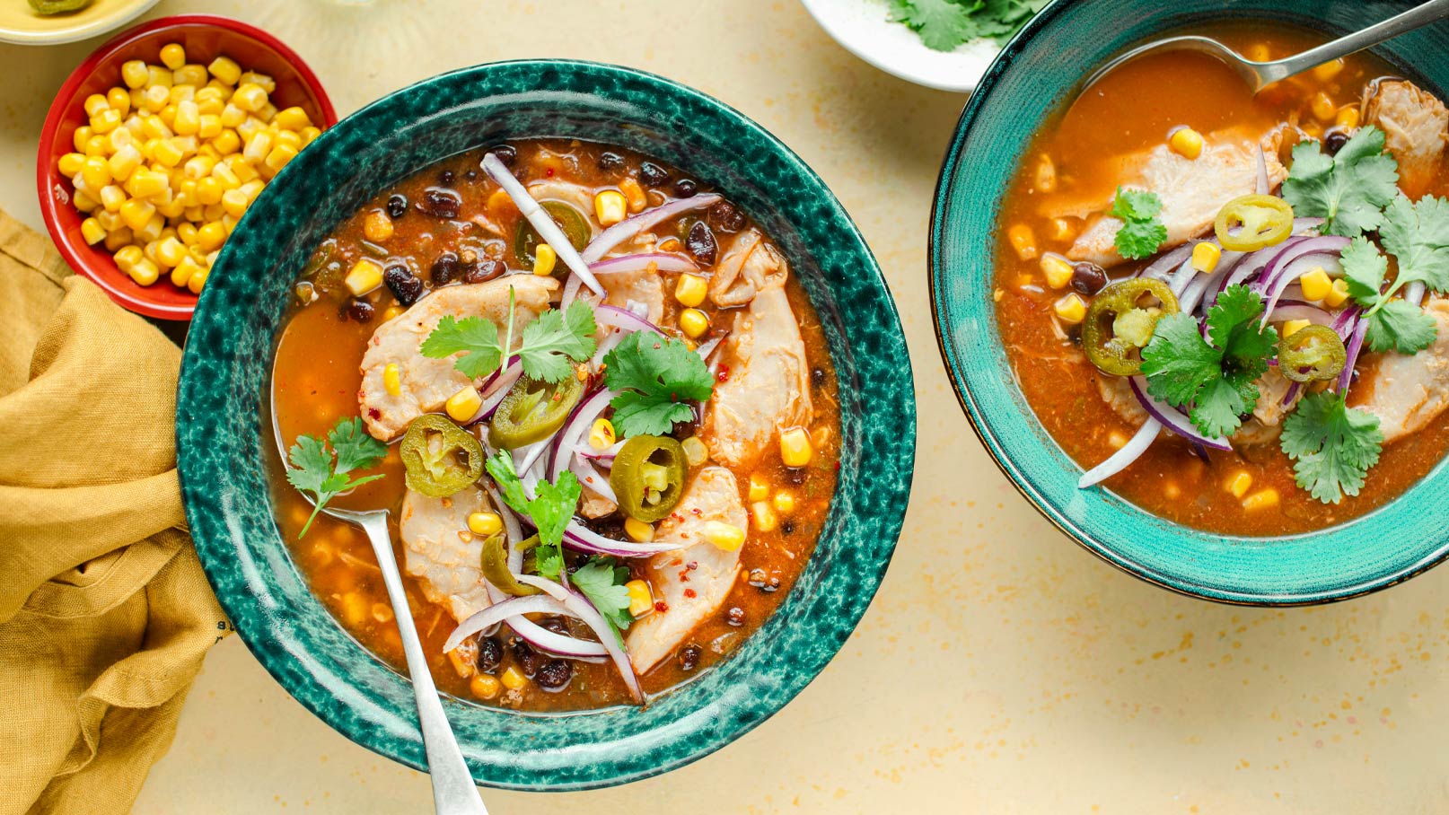 Zwei Schalen mit einer herzhaften Suppe, die Stücke von Hühnerfleisch, schwarze Bohnen, Mais, rote Zwiebeln und Jalapeños enthält. Frische Korianderblätter garnieren die Suppe, die in hellen, geflammten Schalen serviert wird. Neben der Suppe steht eine Schale mit Maiskernen.
