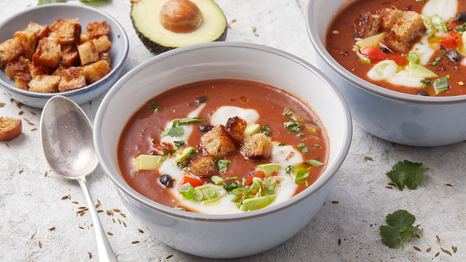 Eine Schüssel mit Tomatensuppe, garniert mit Sauerrahm, frischen Kräutern, gewürfeltem Gemüse und Croûtons. Neben der Suppe liegt eine silberne Löffel. Im Hintergrund ist eine kleine Schüssel mit zusätzlichen Croûtons und eine halbierte Avocado sichtbar.