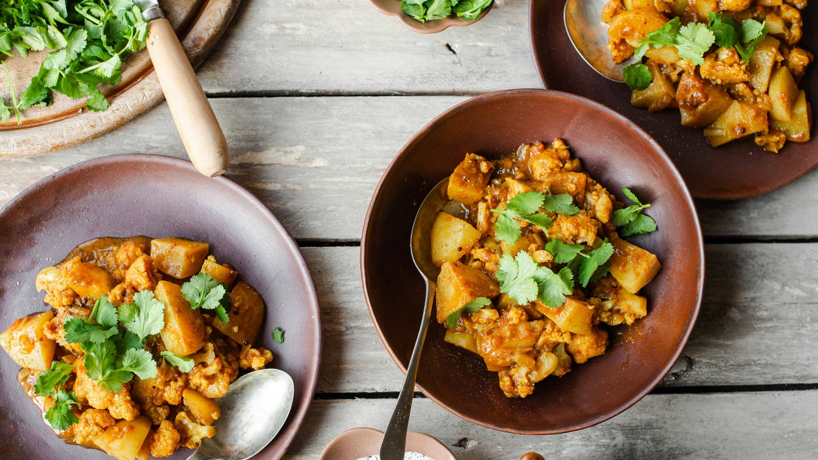 Drei tiefe Schalen mit einem bunten Gericht aus Blumenkohl und Kartoffeln, garniert mit frischem Koriander. Das Gericht ist in einem aromatischen, roten Curry-Sud zubereitet und steht auf einem rustikalen Holztisch.