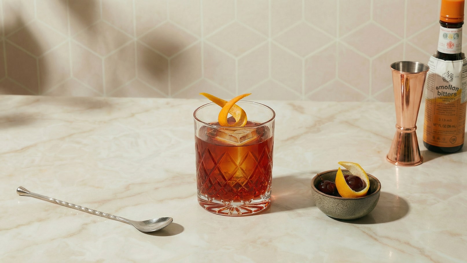 Ein Glas mit einem dunkelroten Cocktail steht auf einem marmorierten Tisch. Der Drink ist mit einer gelochten Eiswürfel und einer spiralförmigen Zitronenschale garniert. Neben dem Glas befindet sich eine kleine Schale mit Kirschen und einer weiteren Zitronenschale.