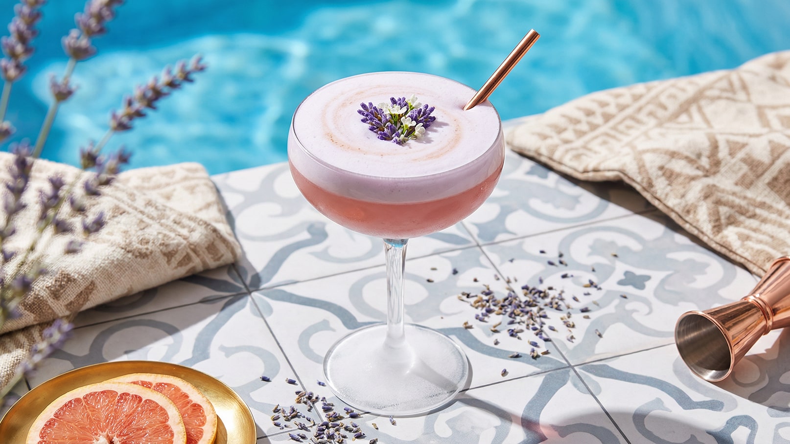 Ein eleganter Cocktail in einer Glasschale, mit einer lila Schaumhaube und dekorativen Essenzen von Lavendel. Er steht auf einem dekorativen Tisch, umgeben von Lavendelblüten und einer gefalteten Decke, während im Hintergrund ein blauer Pool sichtbar ist.