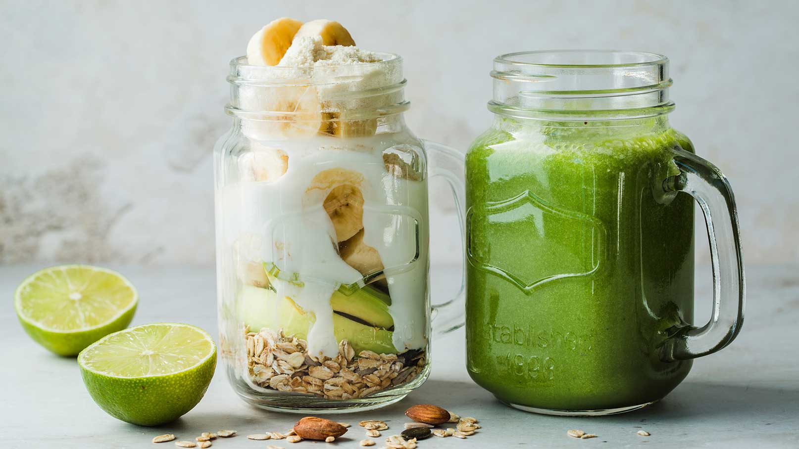 Das Bild zeigt zwei Gläser: Links ein Glas mit einer Schicht aus Haferflocken, Joghurt, Bananen und Apfelstücken. Rechts ein grüner Smoothie in einem Glas. Daneben liegen einige halbierte Limetten.