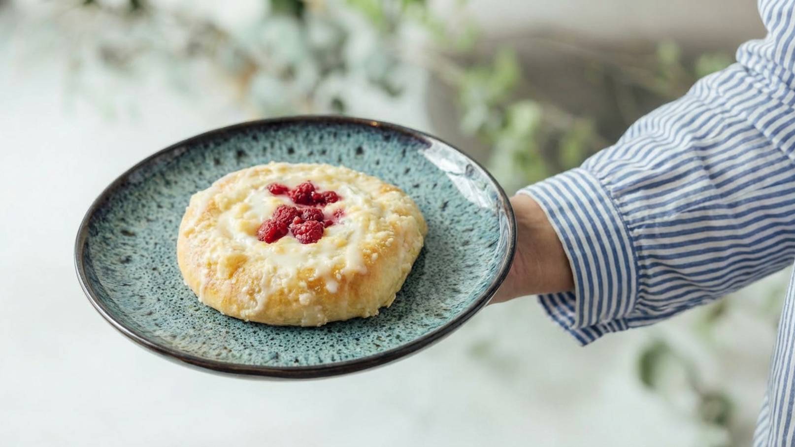 Eine Person hält einen Teller mit einem Gebäck. Das Gebäck hat eine goldene Farbe und ist mit einer cremigen Füllung und frischen Himbeeren dekoriert. Der Teller ist blaugrün und hat einen schlichten, eleganten Rand.
