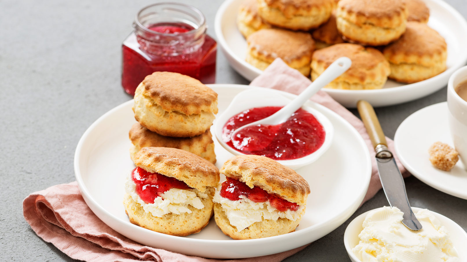 Auf dem Bild sind frisch gebackene Scones auf einem Teller zu sehen, gefüllt mit Sahne und Erdbeermarmelade. Daneben befindet sich eine Schale mit weiterer Marmelade und ein Glas mit Marmelade im Hintergrund. Der Tisch ist in neutralen Tönen gehalten.