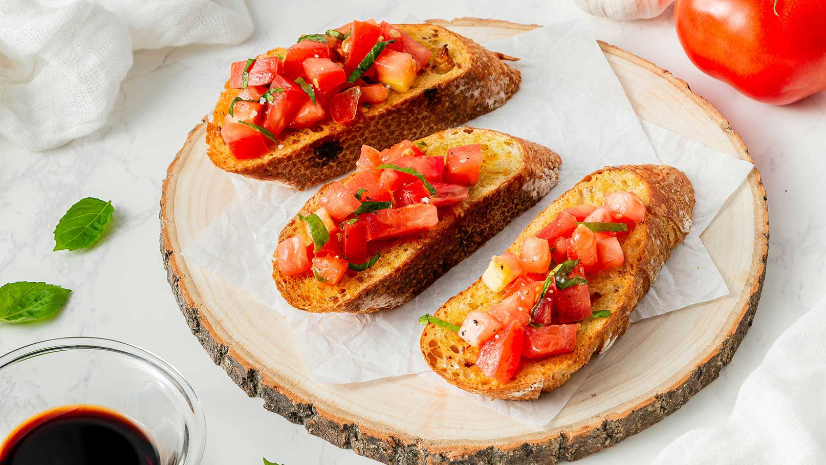 Drei Scheiben geröstetes Brot auf einem Holzbrett, belegt mit einer bunten Mischung aus gewürfelten Tomaten und frischen Kräutern. Ein kleiner Schälchen mit dunkler Flüssigkeit und einige Blätter frischer Minze sind ebenfalls sichtbar.