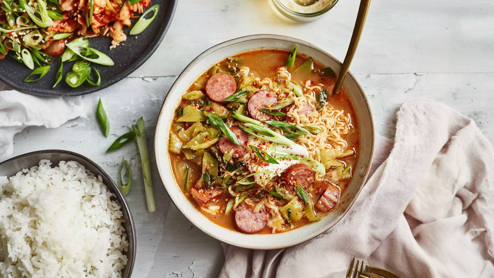 Eine Schüssel mit scharfer Nudelsuppe, gefüllt mit Tofu, Gemüse, Pilzen und frischen Kräutern. Die Suppe hat eine rötliche Farbe und ist mit Glasnudeln garniert. Neben der Schüssel stehen kleine Schalen mit verschiedenen Beilagen.