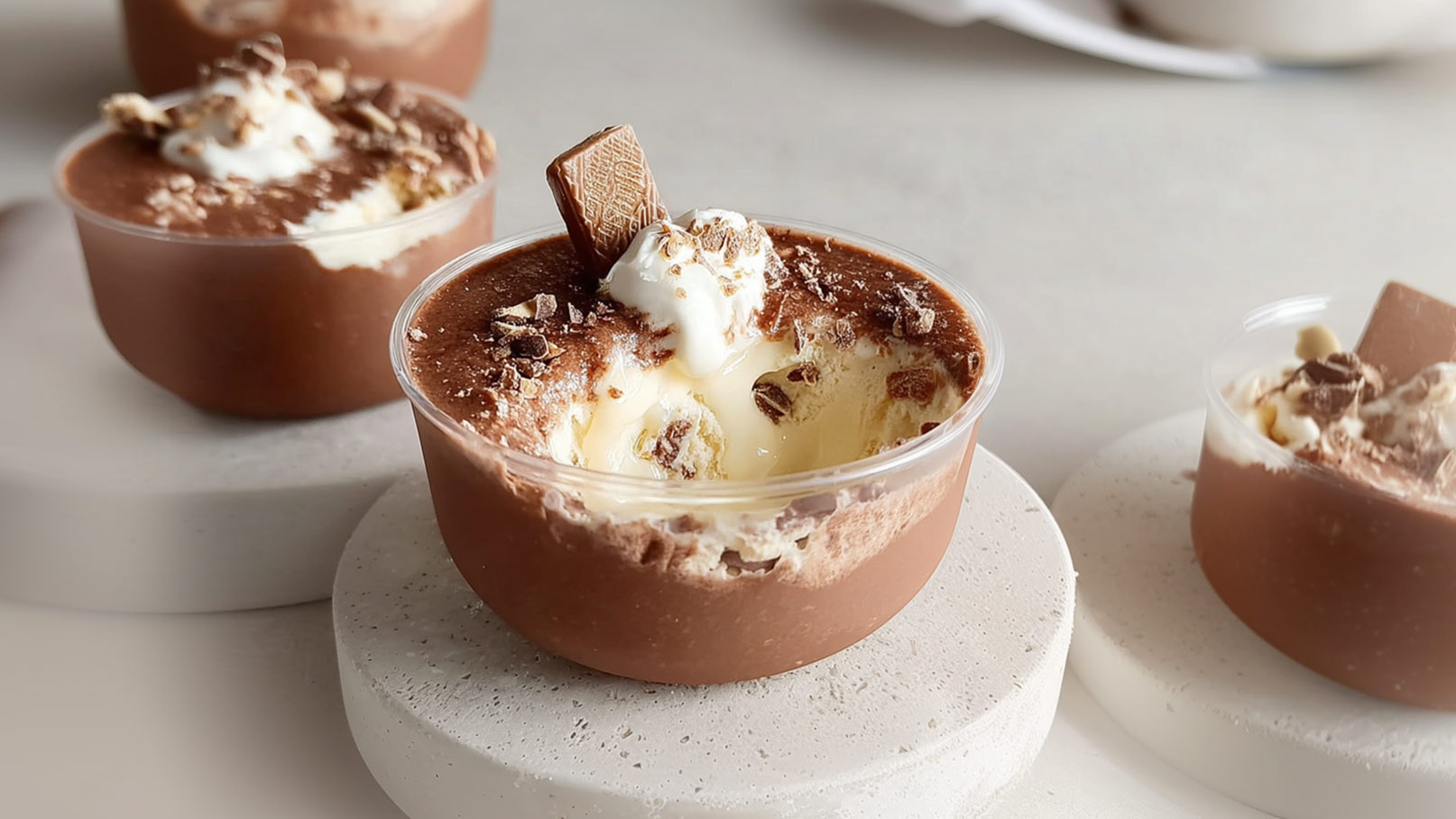 Ein Becher mit schokoladigem Dessert, dekoriert mit Sahne und Schokoladenstückchen. Die Oberfläche ist cremig und hat eine feine Schicht aus Schokoraspeln. Im Hintergrund sind ähnliche Desserts sichtbar, die auf hellen Unterlagen stehen.