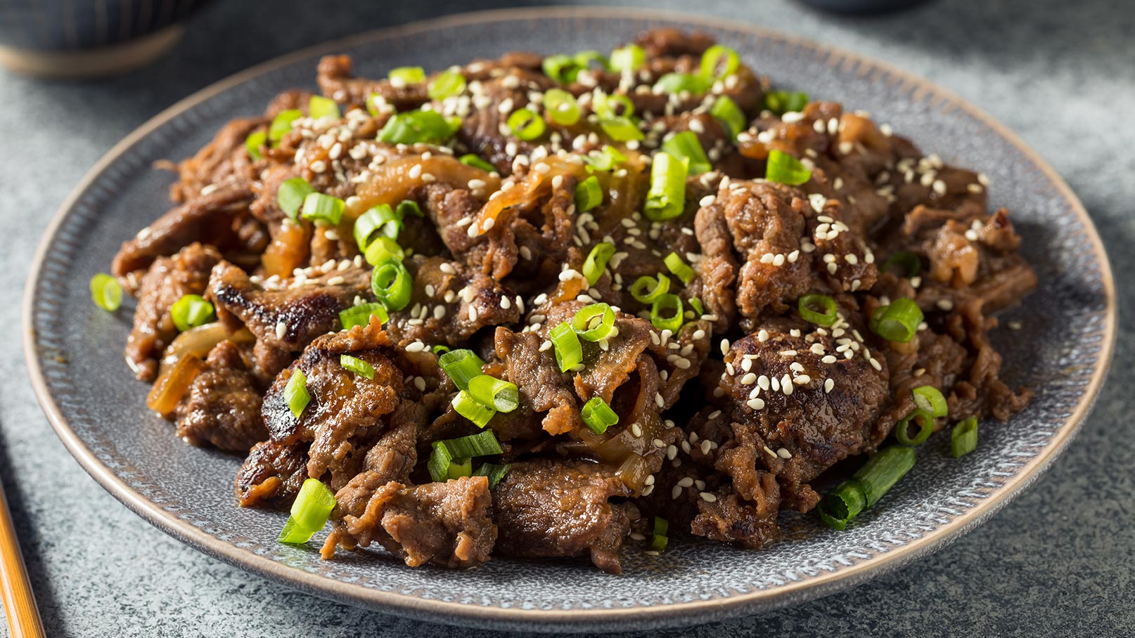 Ein Teller mit gebratenem Fleisch, garniert mit frischen Frühlingszwiebeln und Sesam. Das Gericht wirkt appetitlich und reichhaltig, perfekt für eine asiatische Mahlzeit. Der Teller ist auf einem grauen Untergrund platziert.