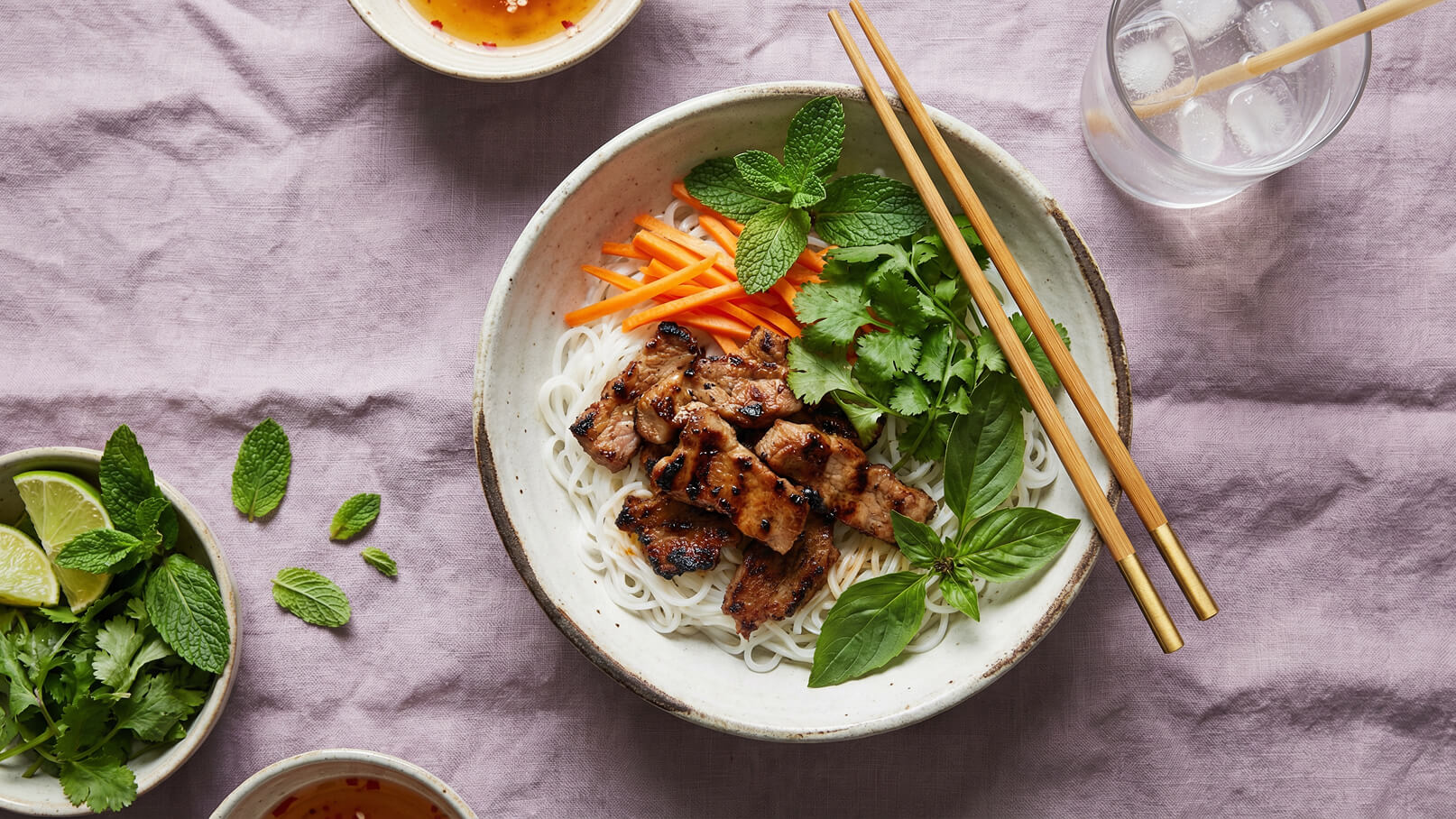 Eine Schüssel mit Reisnudeln, gegrilltem Fleisch, frischen Kräutern, Karottenstreifen und Basilikum, garniert mit Minze. Neben der Schüssel befinden sich Essstäbchen und eine kleine Schale mit Saucen und Limettenscheiben. Der Hintergrund ist ein lila Tuch.