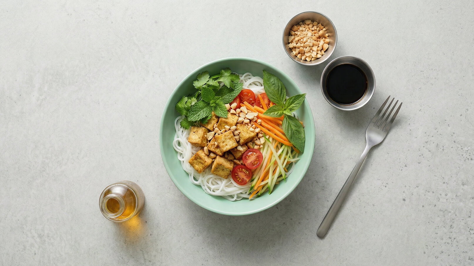 Eine grüne Schüssel enthält Reisnudeln, gewürfelten Tofu, Kirschtomaten, geschnittene Karotten, Gurken und frische Kräuter wie Minze und Basilikum. Daneben befinden sich Schalen mit Erdnüssen und einer dunklen Soße sowie eine Gabel. Der Hintergrund ist hell und einfarbig.