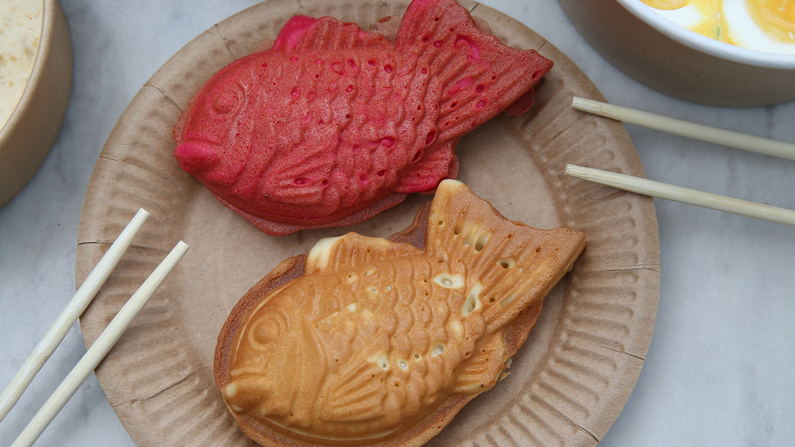 Auf einem Pappteller liegen zwei fischförmige Süßigkeiten: eine rote und eine braune. Sie sind knusprig und leicht gewellt. Daneben befinden sich zwei Essstäbchen, die für die Zubereitung oder den Verzehr verwendet werden können.
