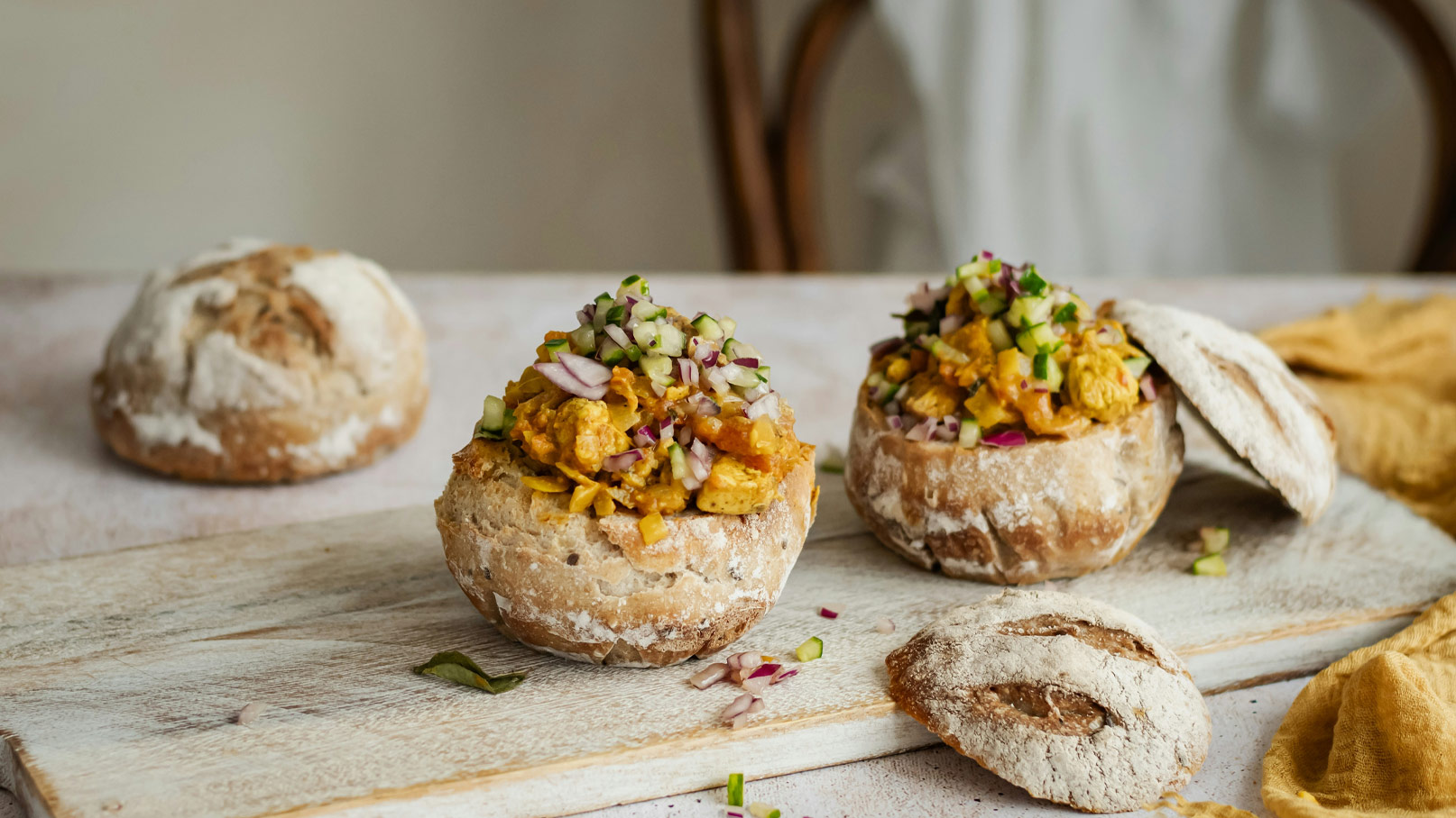 Zwei aufgeschnittene Brötchen auf einem Holzbrett, gefüllt mit bunt gewürfeltem Gemüse und Hühnerfleisch. Eines der Brötchen liegt halb aufgeklappt neben dem anderen. Die Füllung besteht aus leckeren Zutaten, die ansprechend angeordnet sind.