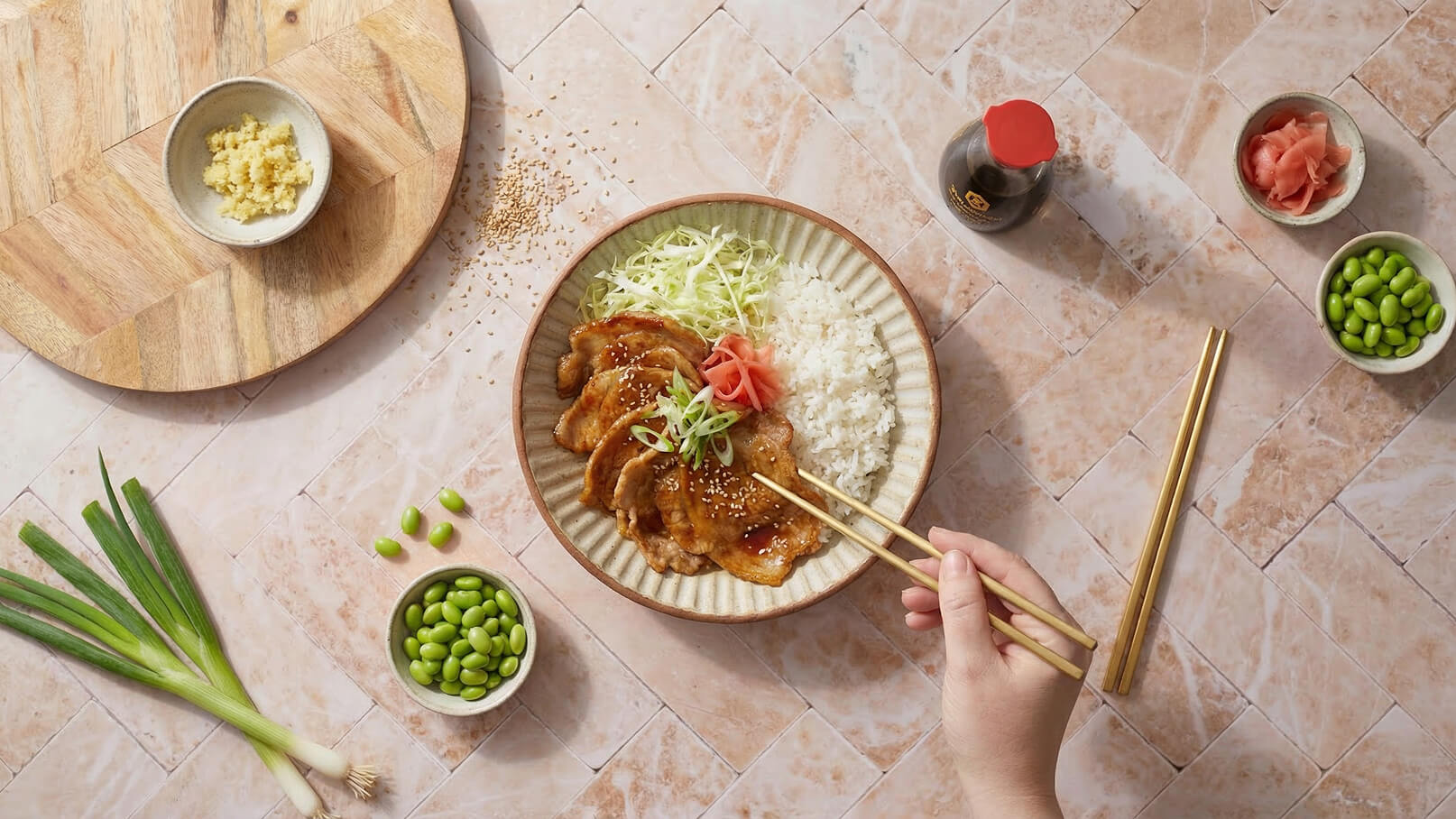 Ein Teller mit gebratenem Fleisch, Reis und frischem Gemüse ist abgebildet. Neben dem Teller sind Schälchen mit Ingwer, Sojasauce und Edamame sichtbar. Eine Hand hält Essstäbchen, bereit, das Essen zu genießen.