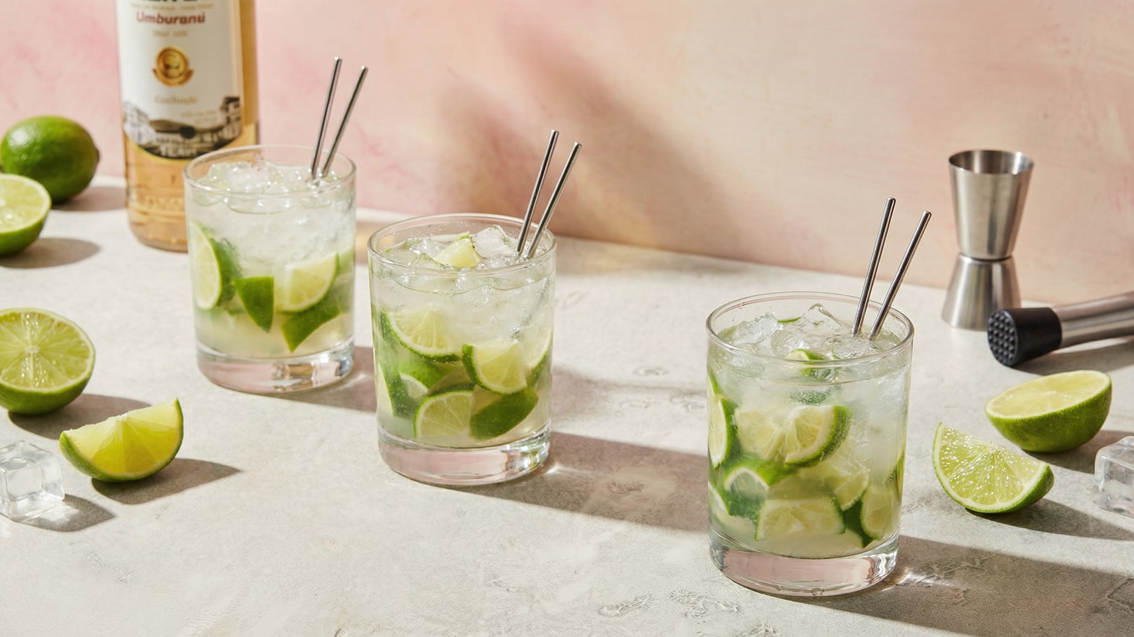 Drei Gläser mit Cocktails, gefüllt mit Eis und Limettenscheiben, stehen auf einer Tischoberfläche. Um die Gläser sind mehrere Limettenhälften und Eiswürfel verteilt. Im Hintergrund ist eine Flasche alkoholischer Getränke sichtbar.