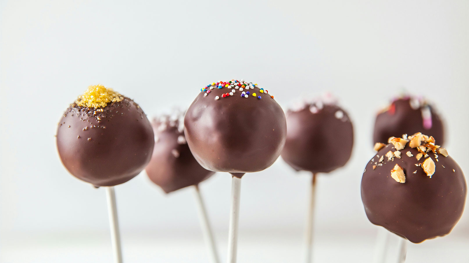 Sechs Schokoladen-Lollis auf weißen Stielen stehen in einer Reihe. Die Lollis sind mit verschiedenen Toppings dekoriert, darunter farbige Streusel, gehackte Nüsse und glitzernder Zucker. Der Fokus liegt auf den glänzenden Schokoladenoberflächen und den bunten Verzierungen.