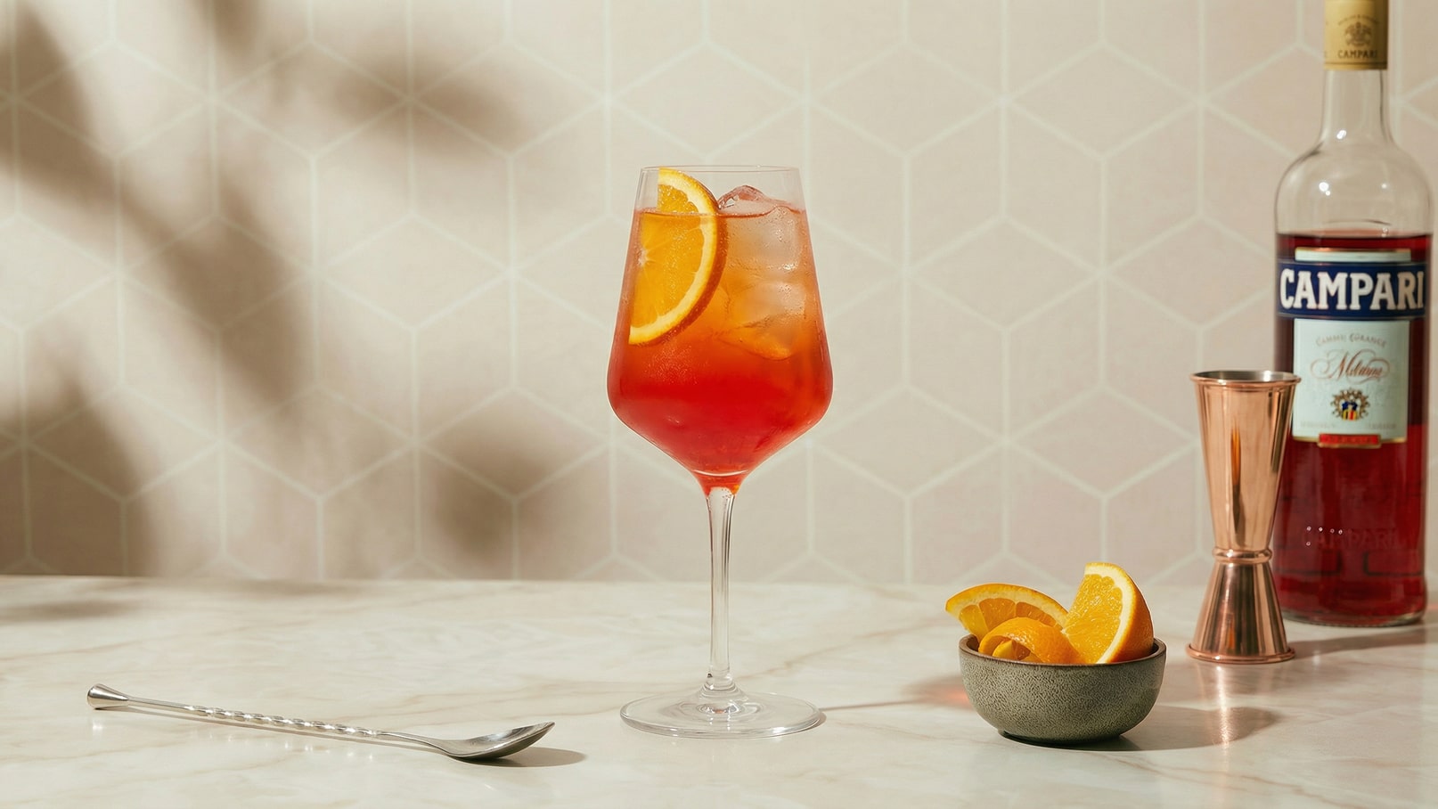 Ein stilvoller Cocktail in einem großen Weinglas, gefüllt mit Eis und Garnituren aus Orangenscheiben. Daneben befindet sich eine kleine Schale mit weiteren Orangenschnitzen und eine Flasche Campari. Der Hintergrund ist dezent mit einem geometrischen Muster gestaltet.