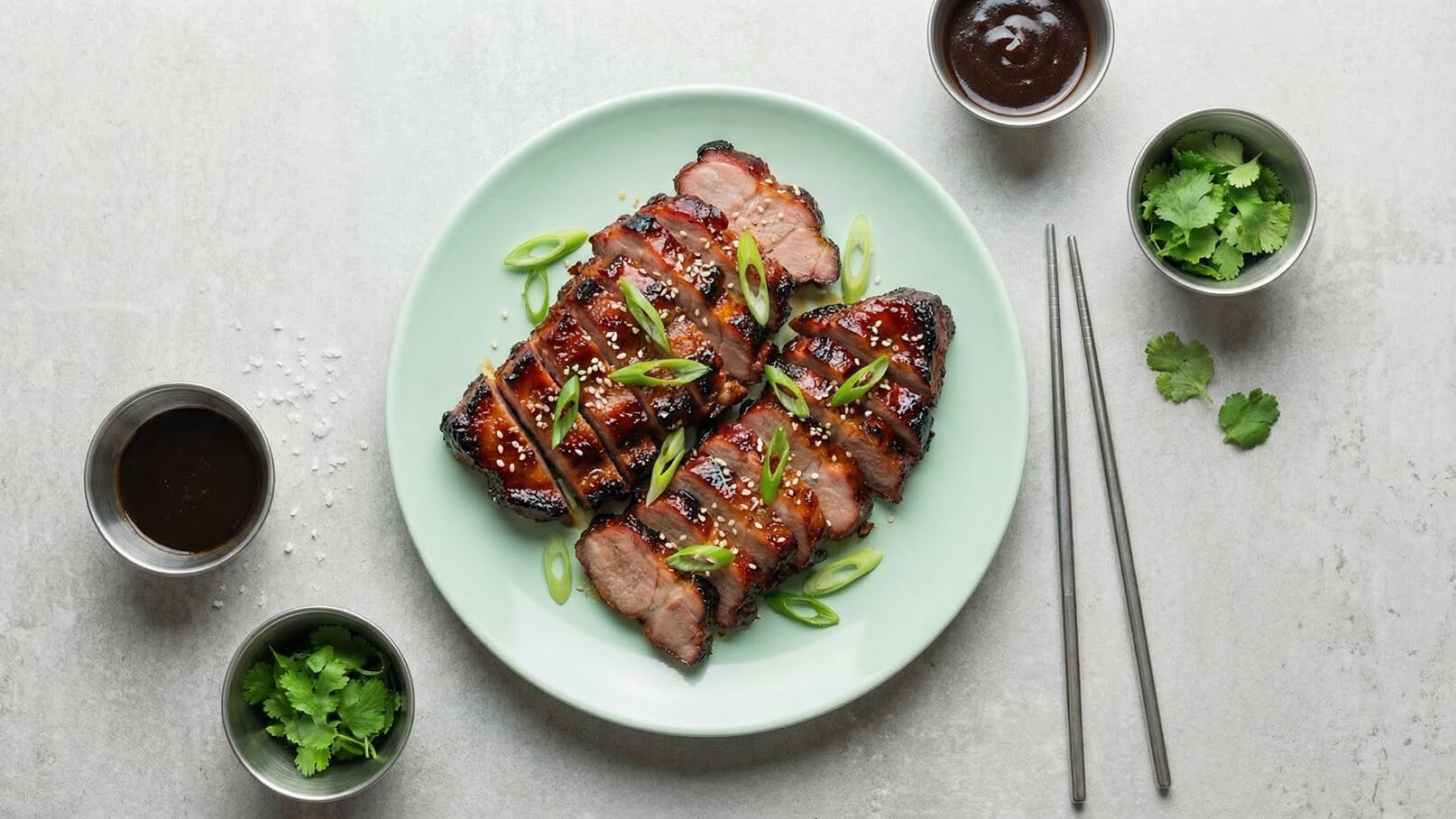 Auf einem grünen Teller liegen zwei Stücke gegrilltes Fleisch, in Scheiben geschnitten und mit Frühlingszwiebeln sowie Sesam bestreut. Daneben befinden sich kleine Schalen mit einer dunklen Sauce und frischem Koriander sowie Essstäbchen.